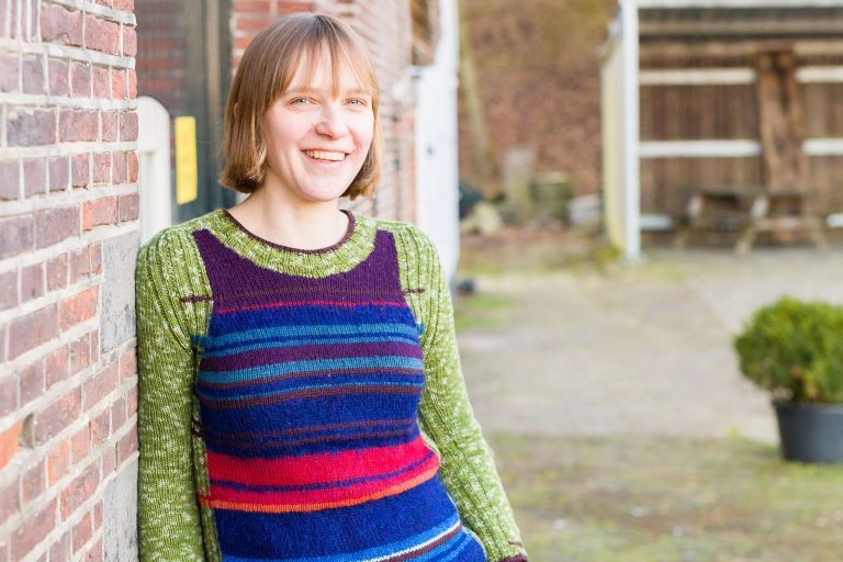Een vrouw staat tegen de muur te leunen. Ze kijkt rechtstreeks de camera in. Ze heeft half blond haar en draagt een gebreide trui die gestreept is met de kleuren groen, blauw, rood, roze en paars.