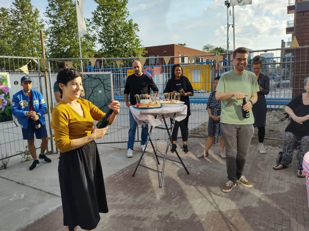 Foto van groep mensen op de opening. Er worden flessen drinken opengemaakt.