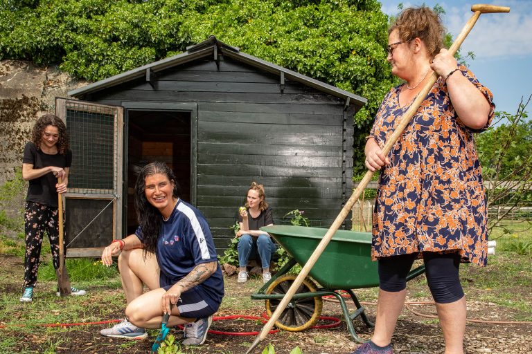 Er staan drie vrouwen die aan het werk zijn in een moestuin. De Linker heeft zwarte kleding aan en houd een schep vast. De middelste heeft blauwe sportkleding aan en kijkt weg met een klein tuinschepje in haar hand. De rechter staat en heeft met beide handen een schoffel in haar handen. Achter haar staat een een groene kruiwagen. Achterin zit een vrouw tegen het moestuinschuurtje die een groene appel aan het eten is.