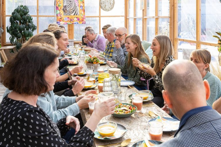 Een groep van ongeveer 20 mensen zitten gezamelijk aan een lange tafel te lunchen en zijn met elkaar in gesprek. De ruimte waarin ze zitten is open en licht.