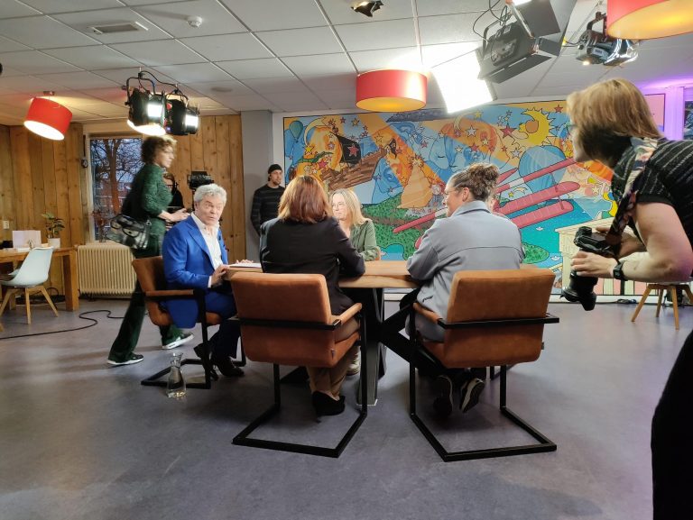 Foto van vijf mensen zitten aan een ovale tafel. Aan de rechterkant zit de tafelhost. Hij heeft een lichtblauw pak aan. Rondom de tafel lopen mensen van de productie.