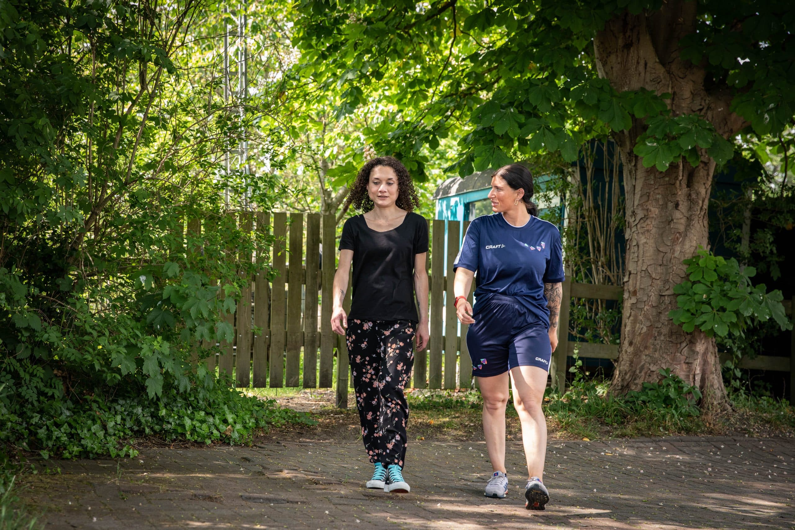 Twee vrouwen lopen buiten naast elkaar. Een vrouw (L) draagt een zwarte broek met bloemetjesprint en de andere vrouw (R) draagt een sporttenue.