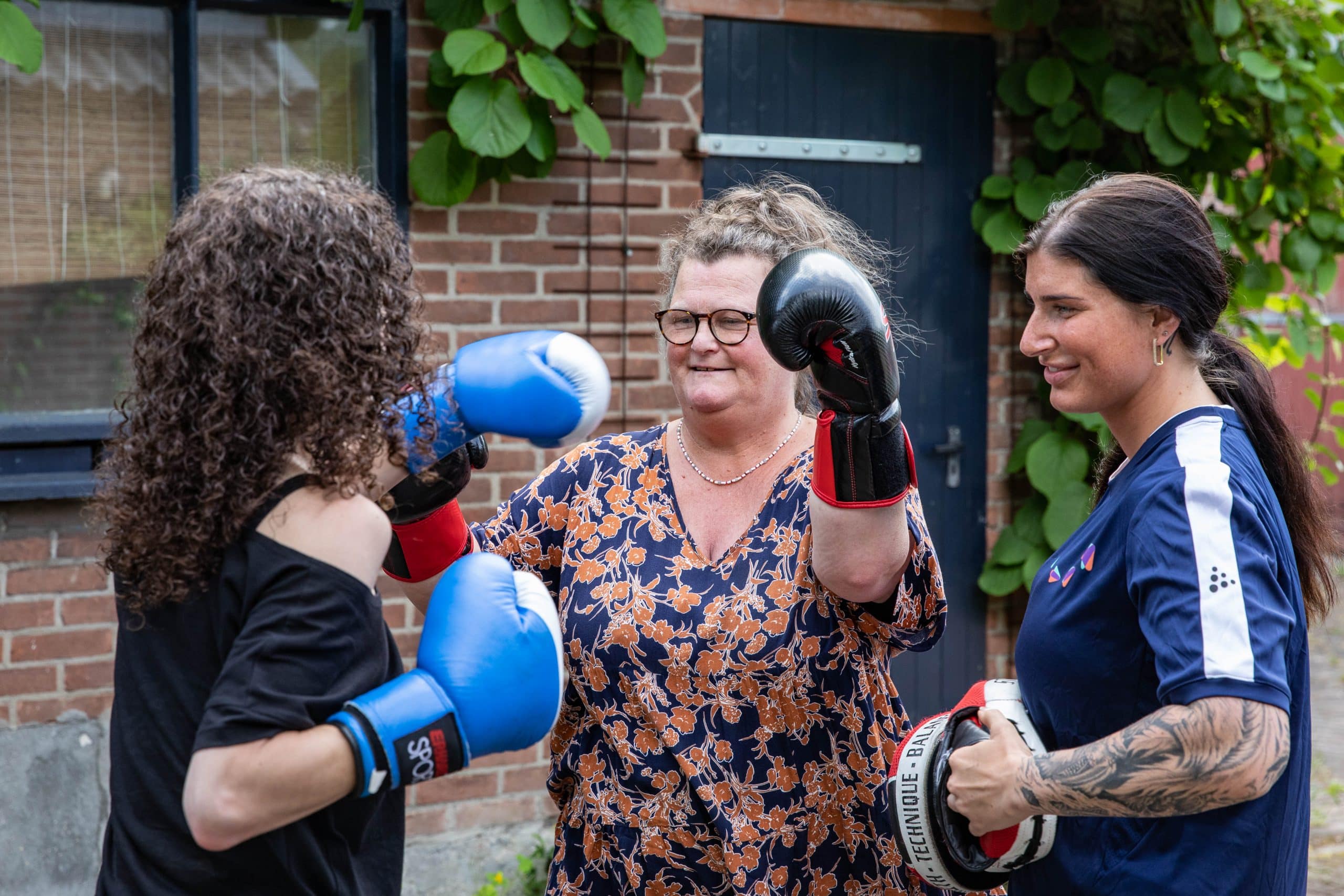 Twee vrouwen zijn aan het boksen, een vrouw draagt een zwart t-shirt, de andere vrouw draagt een oranje-blauw geprint shirt en er staat een trainer bij met een blauw shirt en een getattoeerde arm.