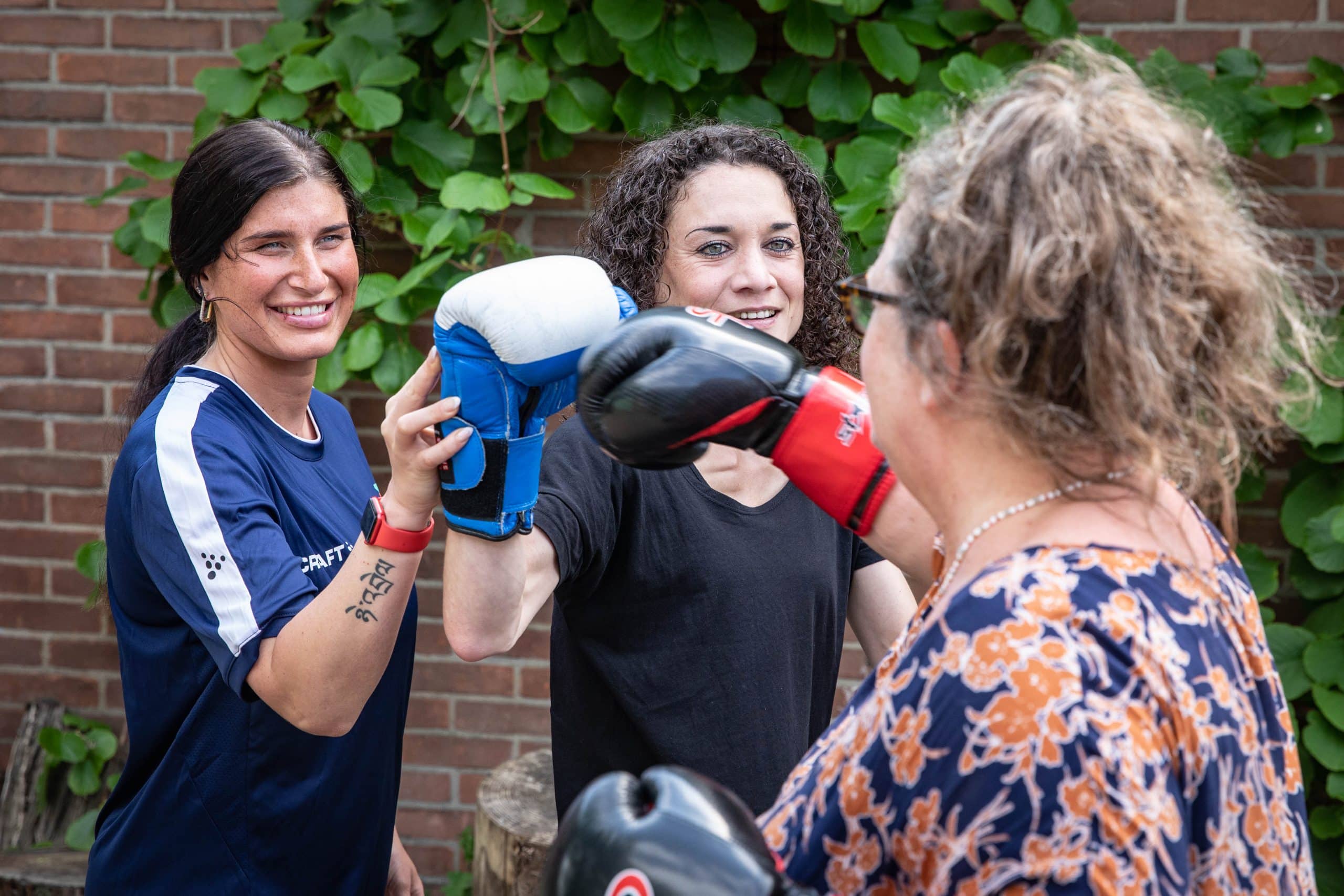 Twee vrouwen hebben beide bokshandschoenen aan en maken een boksbeweging naar elkaar. Een andere vrouw staat erbij en pakt met haar rechterarm de bokshandschoen van de achterste vrouw beet.