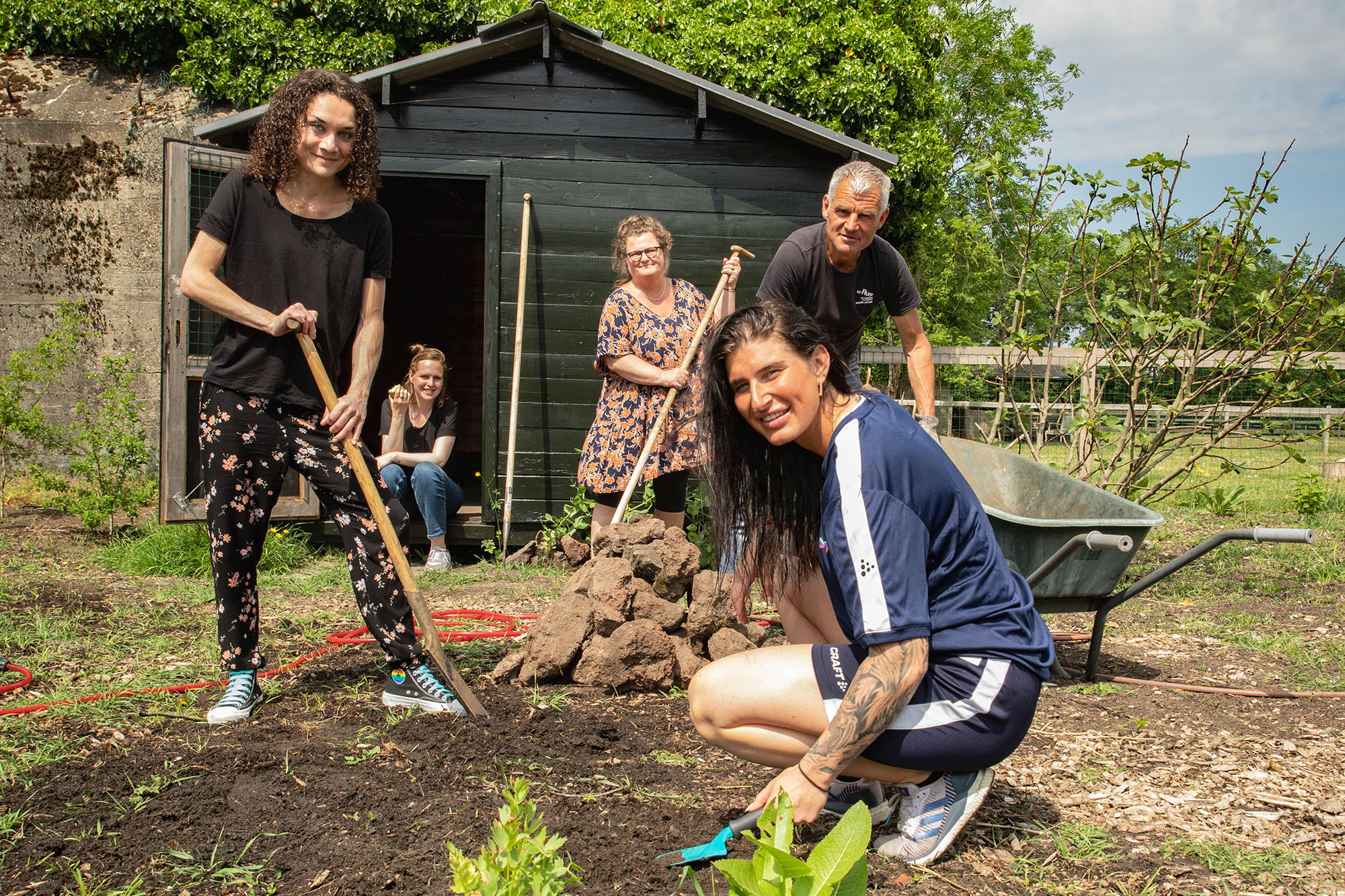Vijf mensen zijn aan het werk in een moestuin. Ze kijken allemaal recht de camera in. Op de achtergrond is een houten tuinhuisje. Aan de rechterkant staat een kruiwagen.