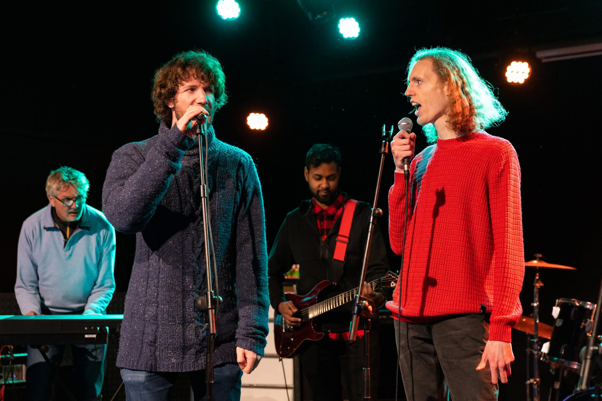 Vier mensen maken muziek op een podium. Één speelt keyboard, een gitaar en twee zingen.