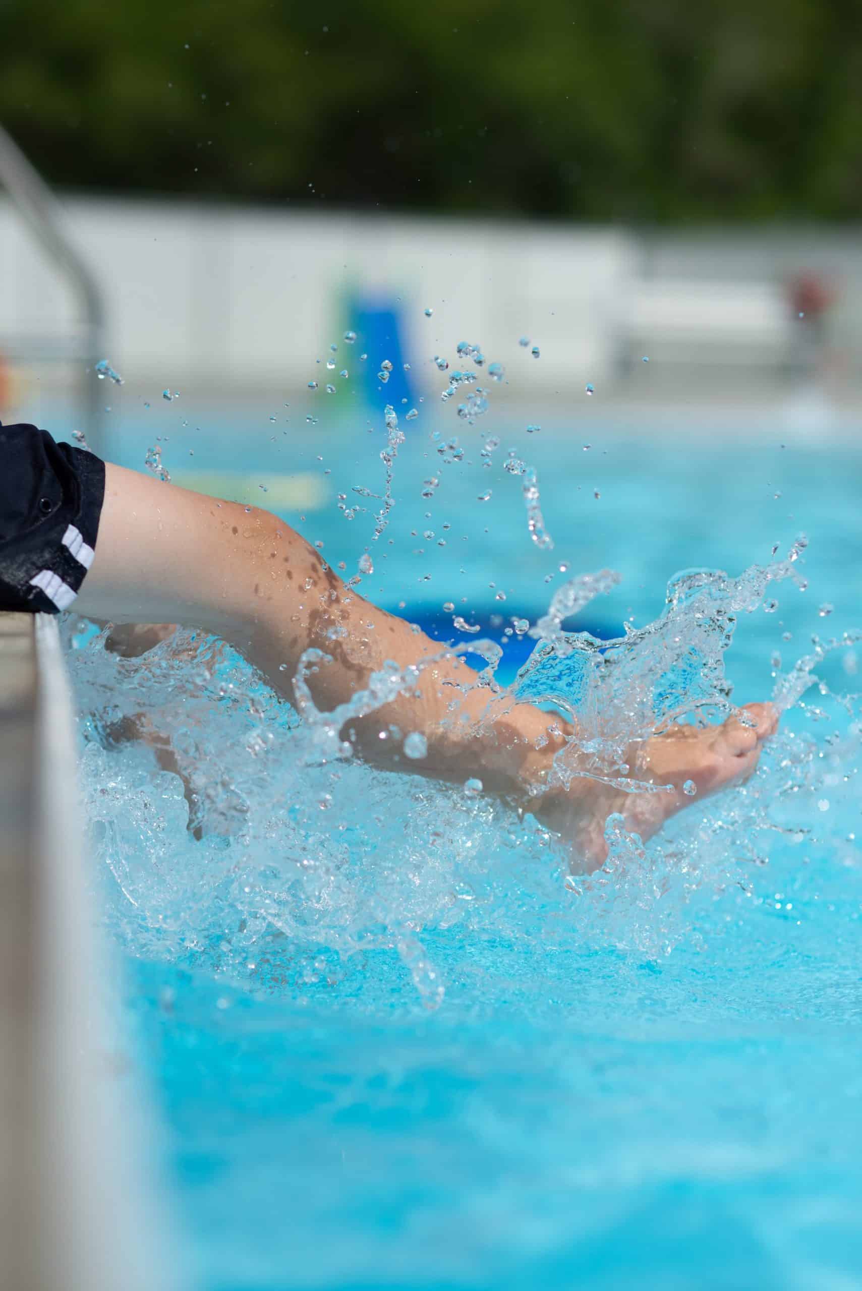 Foto van persoon die op rand van een zwembad zit en met voeten in het water spettert. We zien alleen de onderbenen.